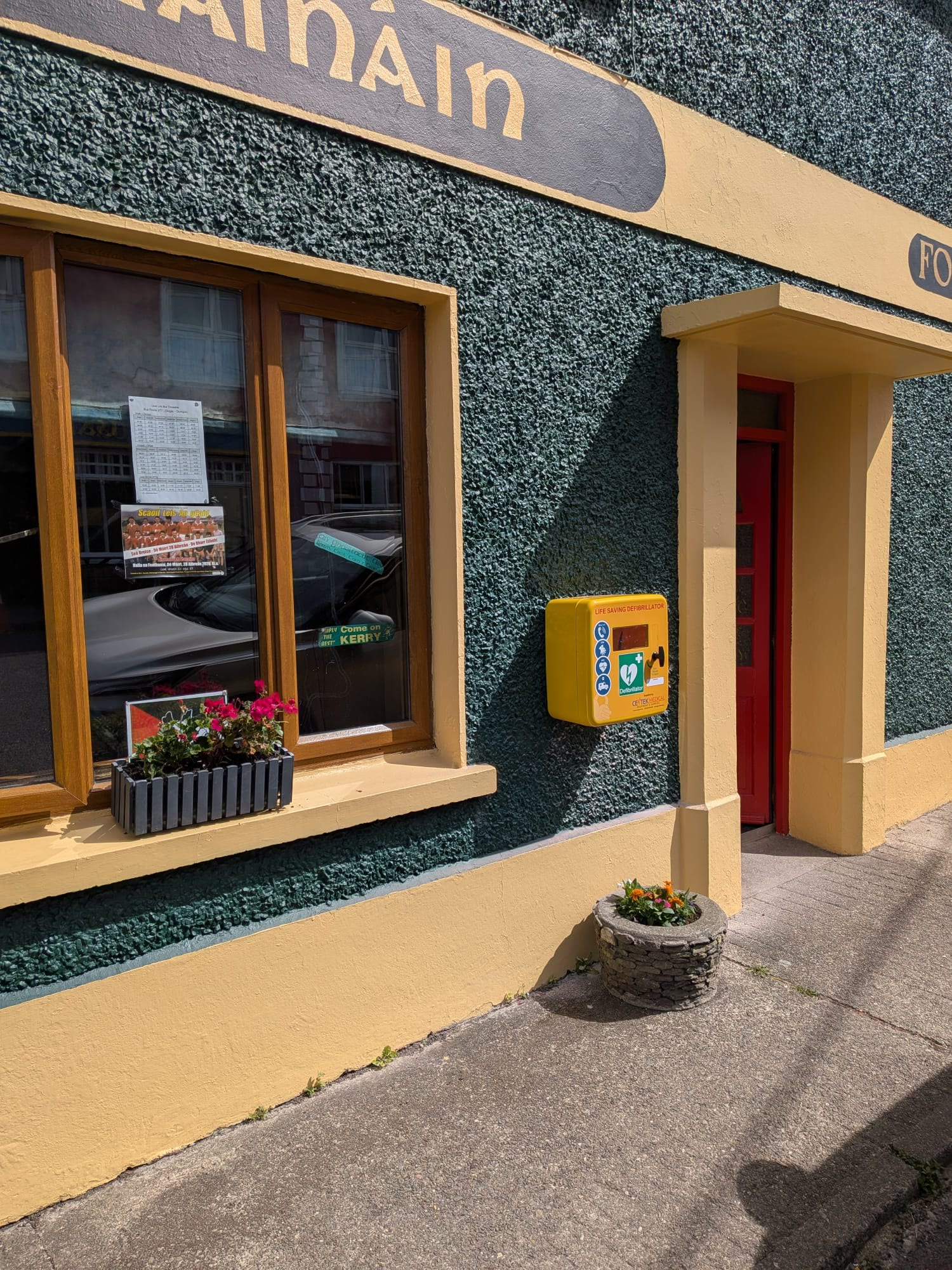 A yellow public-access AED cabinet on the wall of a green pub front, beside a red door