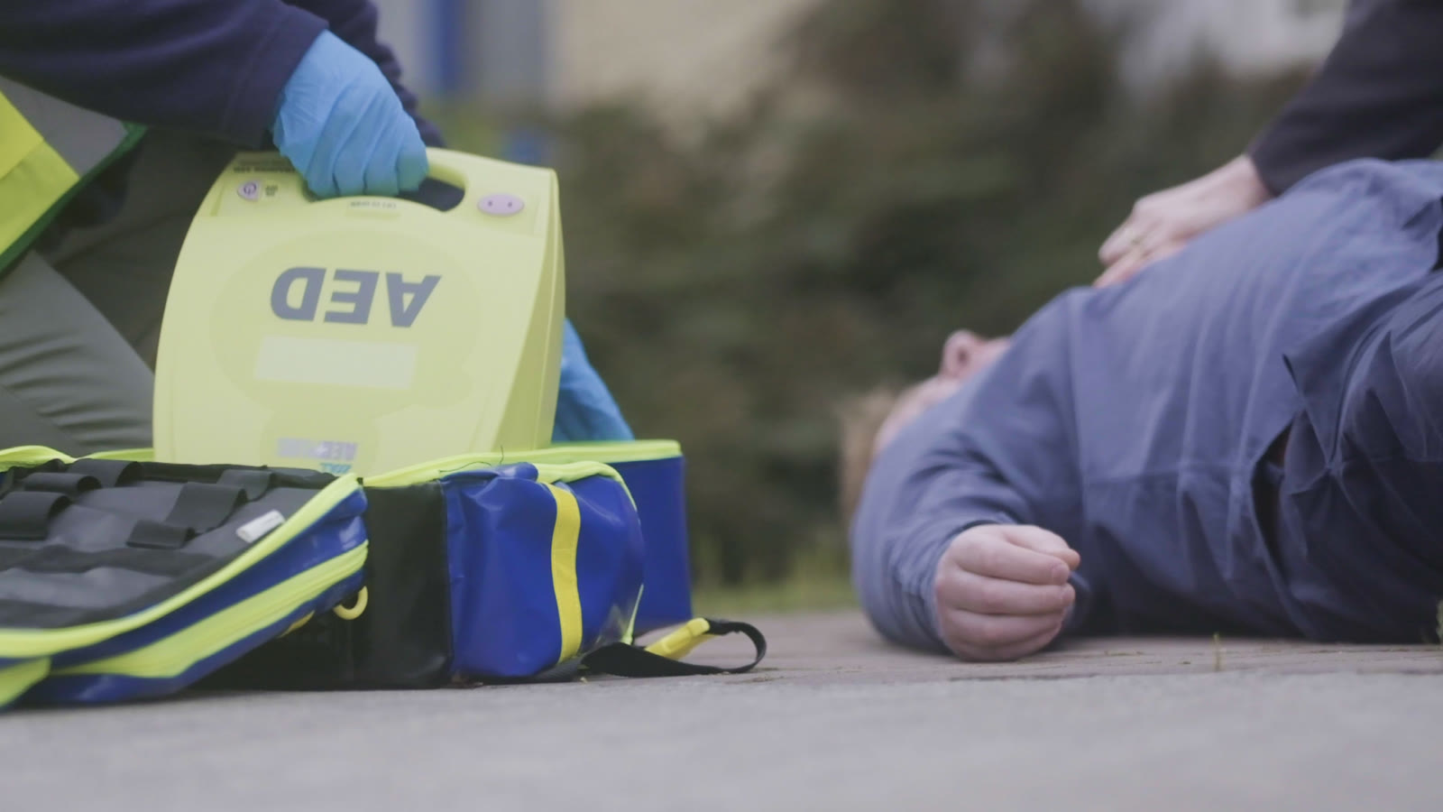 A responder in blue gloves lifts a yellow AED from its response bag beside a person lying on the ground