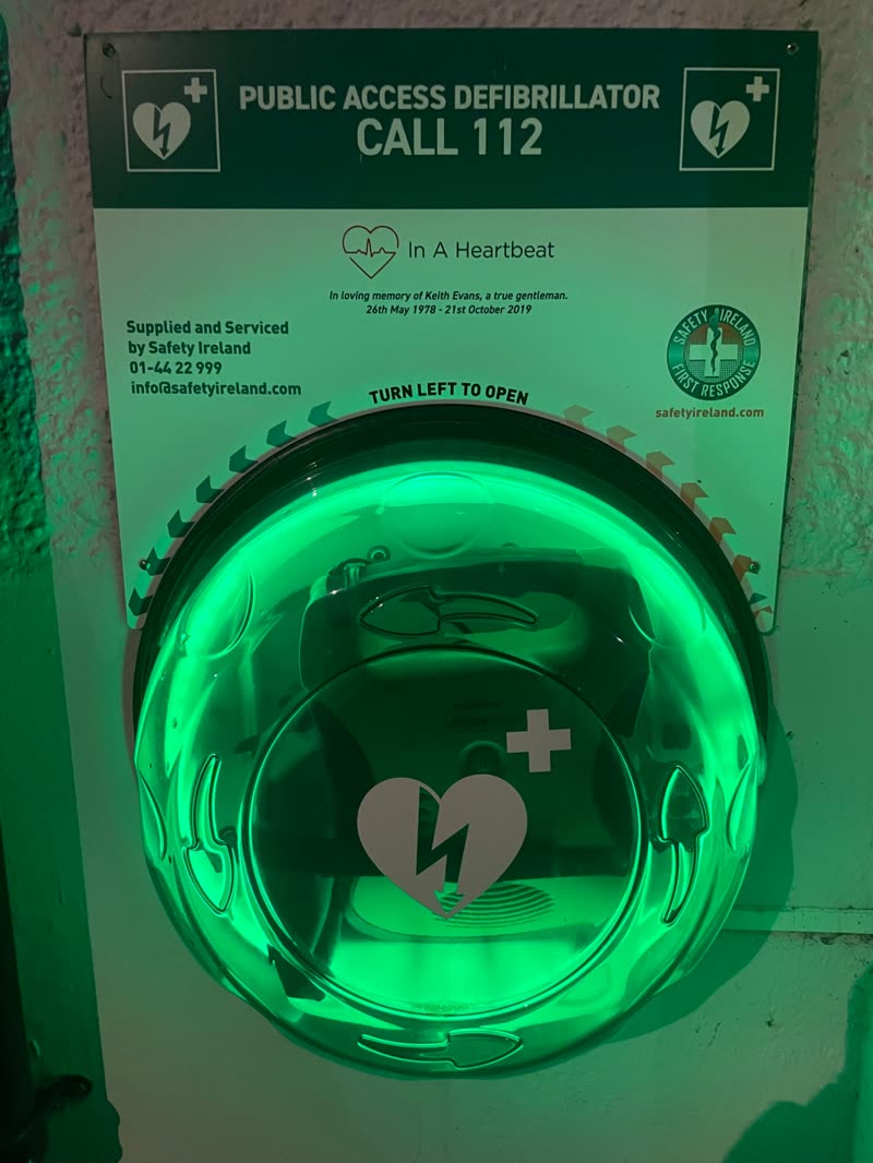 An illuminated green public access defibrillator cabinet on a wall, with a sign reading Call 112