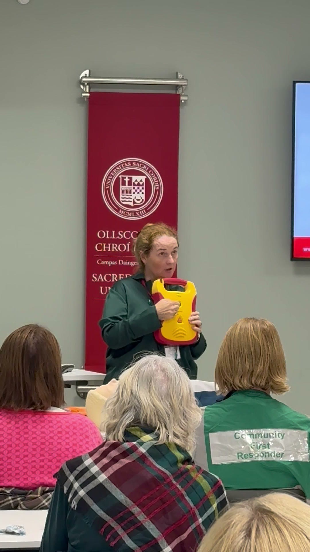 A volunteer holds up a training defibrillator while explaining it to workshop participants